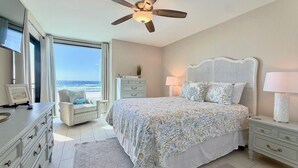 Een slaapkamer met een bed, plafondventilator, dressoir, nachtkastje, lamp, stoel en uitzicht op de oceaan.
