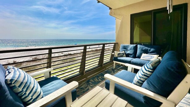 Een balkon met uitzicht op strand en zee, ingericht met blauwe en witte stoelen en een hangende windbel.