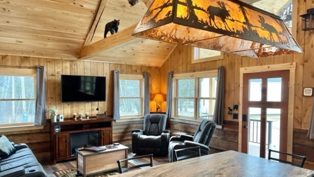 A cozy cabin interior with a wooden interior, a large window, a flat-screen TV, a fireplace, and a dining area. - New Cabin in Eustis.Mtn, River & Dam Views (Eustis)