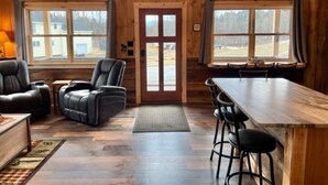 A cozy cabin-style room with a wooden interior, a dining area with a long table, and a large window offering a view of the outside. - New Cabin in Eustis.Mtn, River & Dam Views (Eustis)
