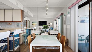 Una cocina moderna con armarios blancos, un comedor con mesa de vidrio y vista al mar a través de un amplio ventanal.