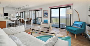 A modern living room with a sofa, a coffee table, and a teal armchair. There is a balcony with a view and a dining area with chairs and a table.