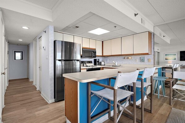 A modern kitchen with a stainless steel refrigerator, microwave, and stove, a bar area with blue chairs, and a dining table with chairs.