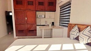 Een slaapkamer met een bed, een houten hoofdbord, een klein tafeltje en een kitchenette met een magnetron en koffiezetapparaat.