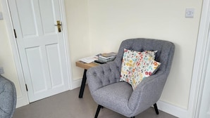 A grey armchair with a floral pillow, a small wooden side table with books, and a white door in a room with light-colored walls and carpet. - 1 Bed Retreat-sleeps 2-secure Parking-garden (Ennis)