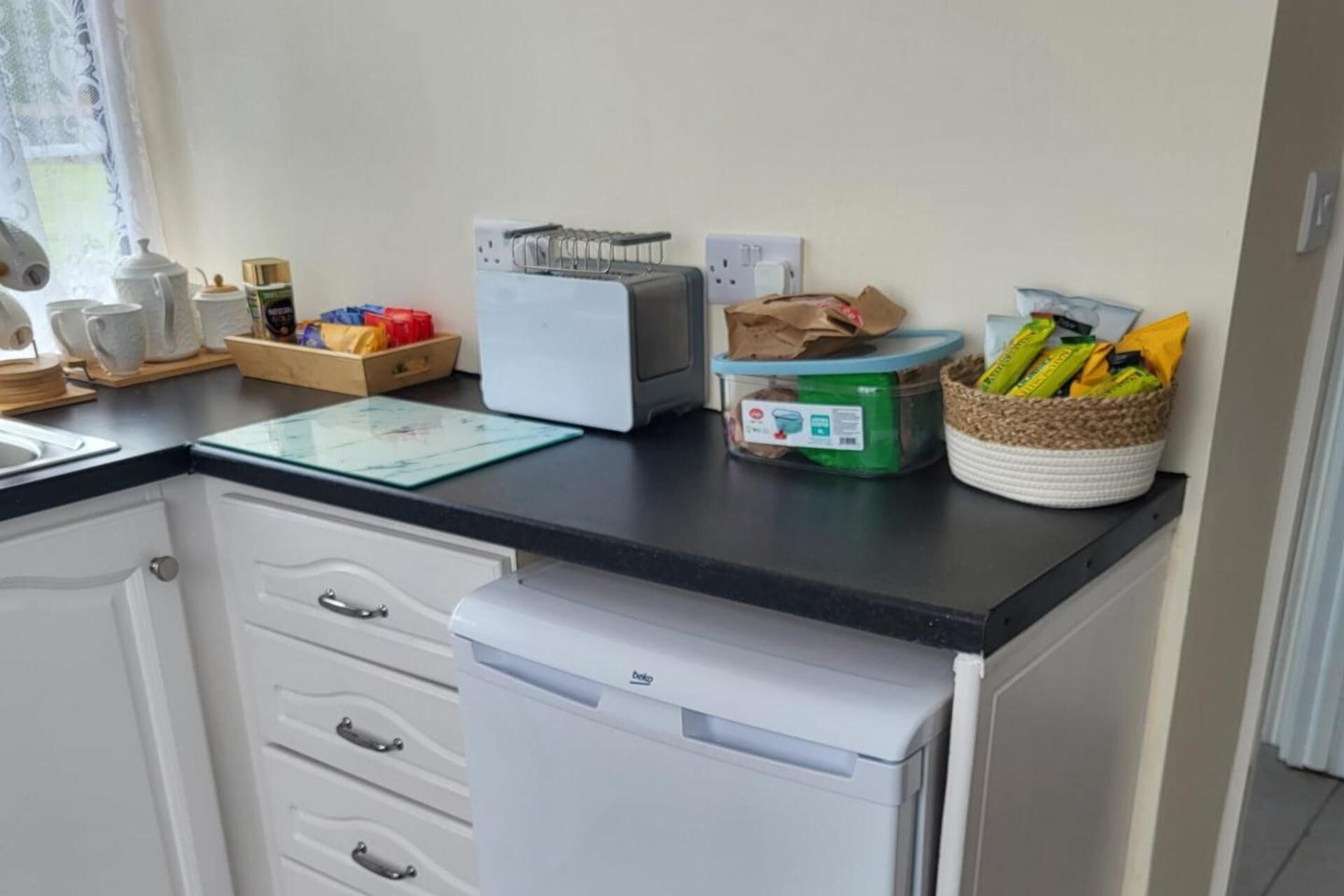 Una cucina con un piano di lavoro nero, mobili bianchi, una macchina per il caffè, un bollitore, un tostapane, un distributore di carta assorbente e un cesto di snack.