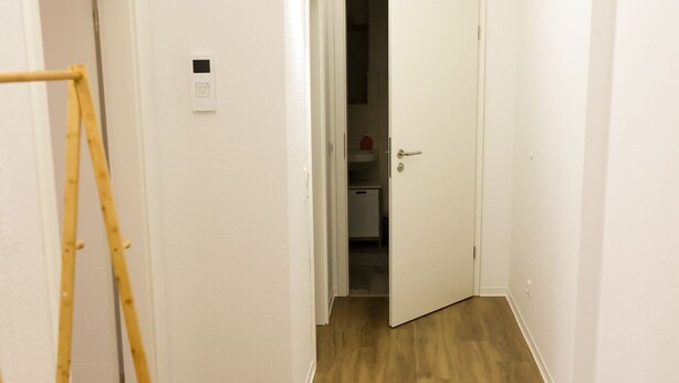Un pasillo con suelo de madera, una puerta blanca y un pequeño cuadro eléctrico blanco en la pared.