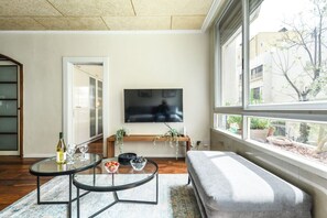 Una sala de estar moderna con un televisor de pantalla plana, una mesa de centro de vidrio, un sofá y un ventanal con vistas a edificios y árboles.