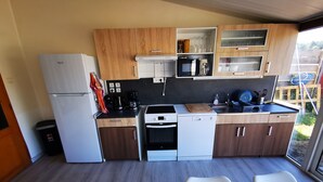 Ett kompakt kök med träskåp, ett vitt kylskåp, en mikrovågsugn, en spis och en diskmaskin. Det finns två vita stolar och ett träbord.
