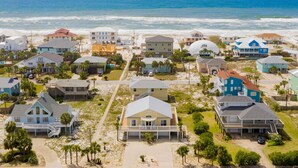 Vue aérienne d’un quartier résidentiel situé sur la côte, avec des maisons, dont certaines donnent directement sur la plage, et une plage en arrière-plan.