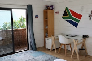 A small, well-lit room with a glass door leading to a balcony, a desk with a chair, a bookshelf, and a South African flag on the wall.