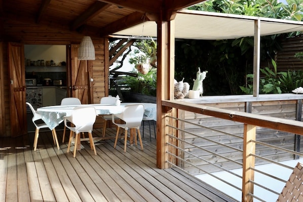A wooden deck with a table and chairs, a hanging light fixture, and a view of greenery.