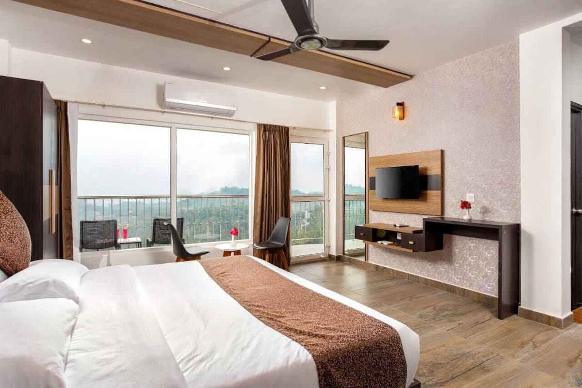 Quarto de hotel com uma cama grande, um ventilador de teto, uma TV fixada na parede, uma mesa e vista da cidade através de grandes janelas.