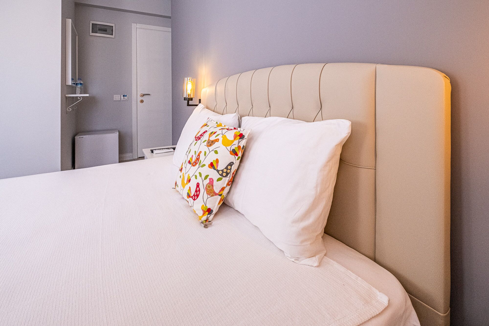 A neatly made bed with a white and beige color scheme, a decorative pillow with a bird pattern, and a wall-mounted light fixture.
