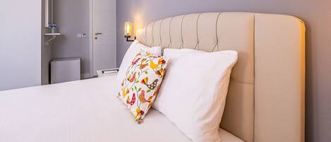 A neatly made bed with a white and beige color scheme, a decorative pillow with a bird pattern, and a wall-mounted light fixture.