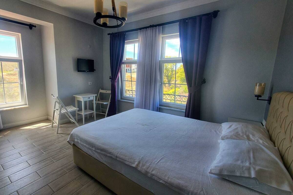 A bedroom with a bed, a desk, and a chair. There are windows with curtains, a TV on the wall, and a chandelier.
