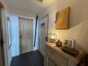 A hallway with a wooden door, a wall-mounted light fixture, a small table with a remote control, a decorative vase, and a microwave.