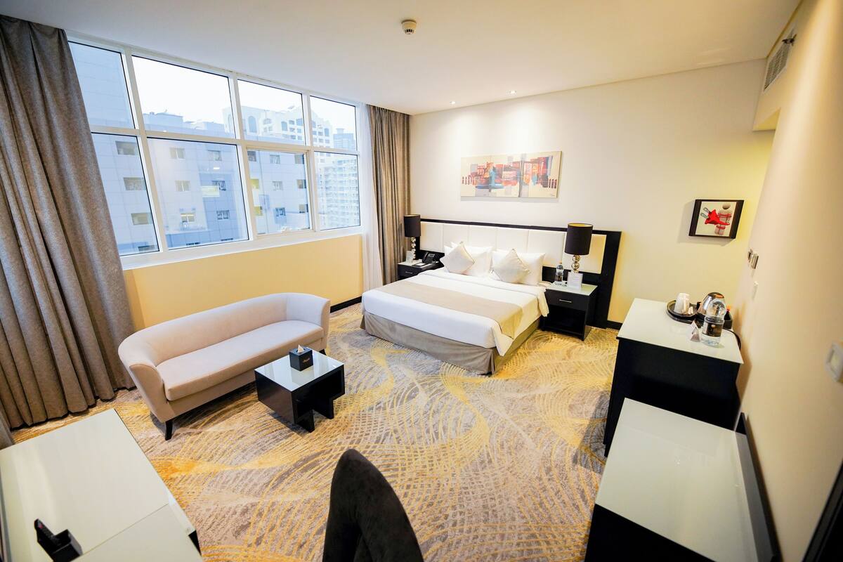 Ein Hotelzimmer mit einem großen Bett, einer Couch, einem Schreibtisch und Blick auf Gebäude durch das Fenster.