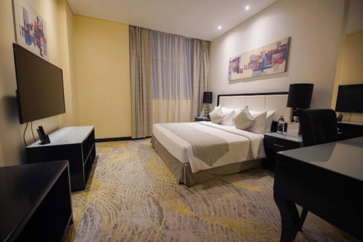 A hotel room with a bed, bedside tables, a desk, a TV, and a painting on the wall.