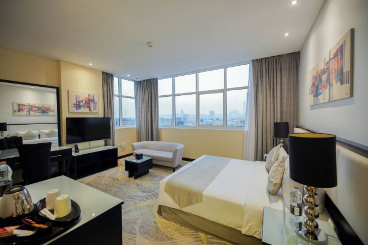 A hotel room with a large bed, a TV, a desk with a chair, a sofa, and a city view through the windows.