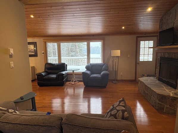 A living room with a fireplace, wooden floors, a stone accent wall, and a view of a snowy landscape through large windows.