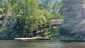 Una propiedad junto al lago con un muelle que se adentra en el agua, rodeada de un bosque denso y una casa en una colina.