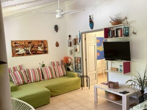 A living room with a green sofa, striped pillows, a flat-screen TV, a ceiling fan, and a bookshelf with books and decorative items.