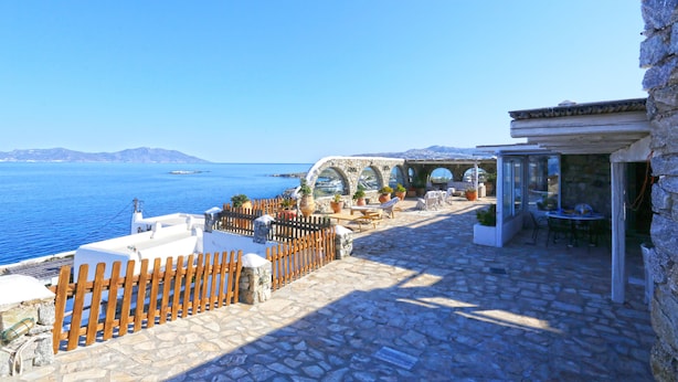 A stone patio with outdoor seating overlooking a body of water, a stone wall, and a clear sky. - Amati Villa Cavo Dellos (Mykonos)