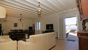 A living room with a sofa, television, dining table, and chairs. There is a door leading to a balcony with a view. - Amati Villa Cavo Dellos (Mykonos)