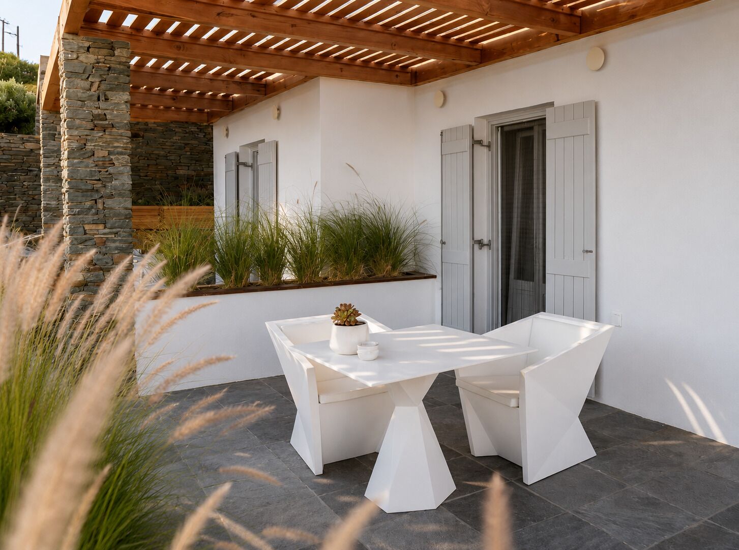 Een modern terras buiten met een witte tafel en stoelen, een stenen pilaar en een houten pergola.