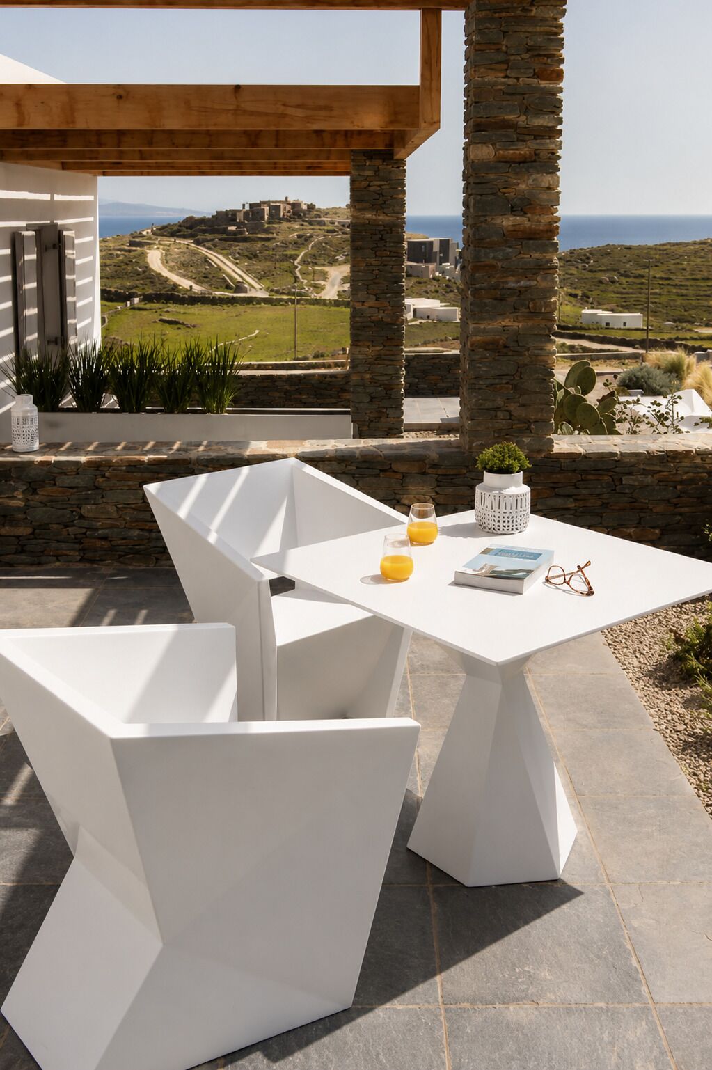 A white table with chairs, a glass of orange juice, a book, and a pair of glasses on a patio with stone pillars and a view of a coastal landscape.