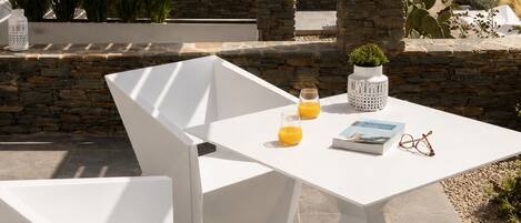 A white table with chairs, a glass of orange juice, a book, and a pair of glasses on a patio with stone pillars and a view of a coastal landscape.