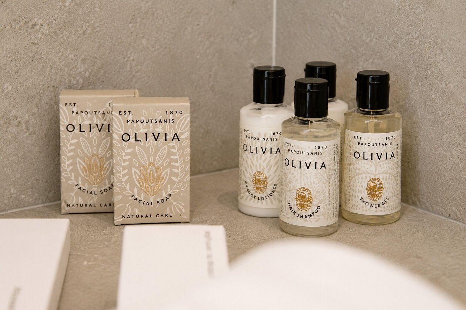 A bathroom with a beige textured wall, a white sink, and four bottles of Olivia brand personal care products.