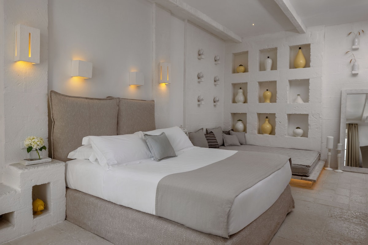 Ein Schlafzimmer mit einem großen Bett, Nachttischen, einem an der Wand befestigten Regal mit Dekoration und einem Spiegel.