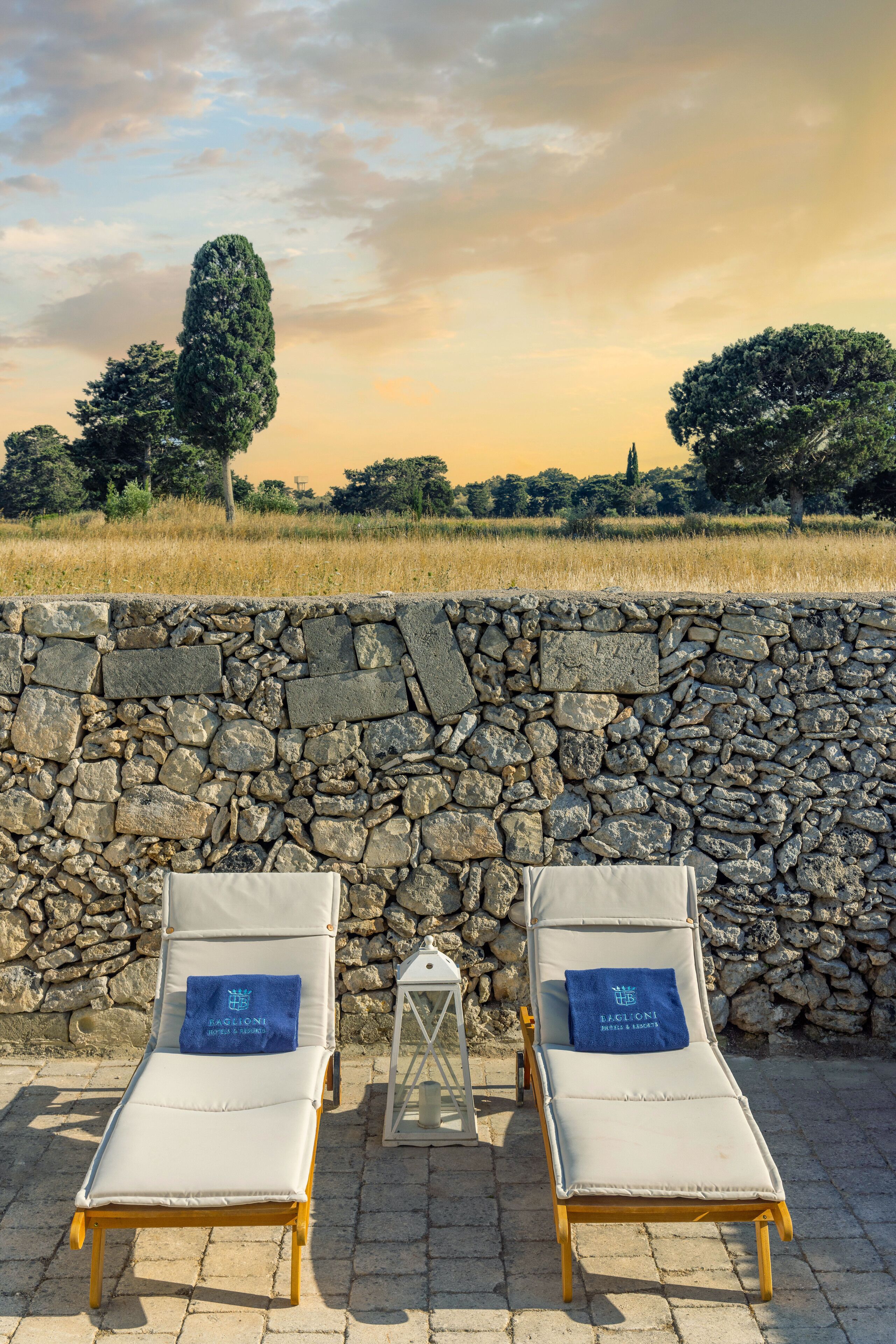 Deux chaises longues avec des serviettes bleues, placées devant un mur de pierre, d’où l’on a vue sur un champ et des arbres.