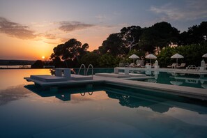 Une piscine à débordement bordée de chaises longues et d’ombrelles, dont l’eau reflète le coucher de soleil et les arbres qui se dressent à l’arrière-plan.