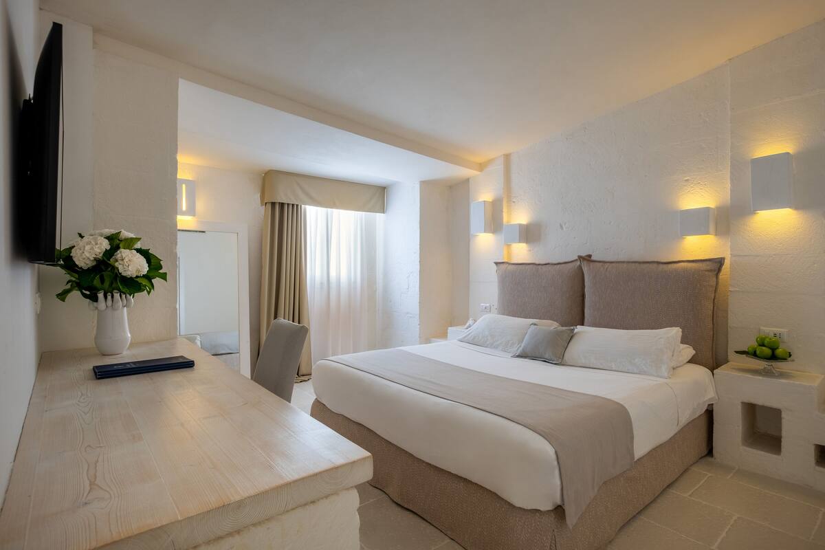 Ein Hotelzimmer mit einem großen Bett, einem Nachttisch mit Blumenvase, einem Flachbildfernseher und Wandleuchten.