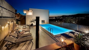 Un espace piscine sur le toit, avec des chaises longues, une petite table et une vue nocturne sur la ville.