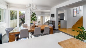 Un espace de salle à manger moderne avec une grande table, des chaises et un lustre. Un escalier mène à un autre niveau, et une plante est placée au premier plan.