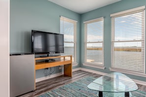 Uma sala de estar moderna com uma televisão de tela plana sobre um suporte de madeira, uma mesa de centro de vidro e janelas amplas com persianas.
