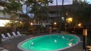 Uma piscina com espreguiçadeiras e guarda-sóis, rodeada por palmeiras e um edifício com varandas.