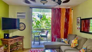 Sala de estar com televisão, relógio, ventilador de teto, sofá com almofadas coloridas e vista para o verde lá fora.