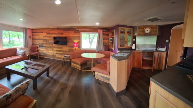 A cozy living area with a sofa, coffee table, and a small round table. There is a wooden wall with a TV mounted above it. - The Terre de feu mobile home (Châteauneuf-sur-Isère)