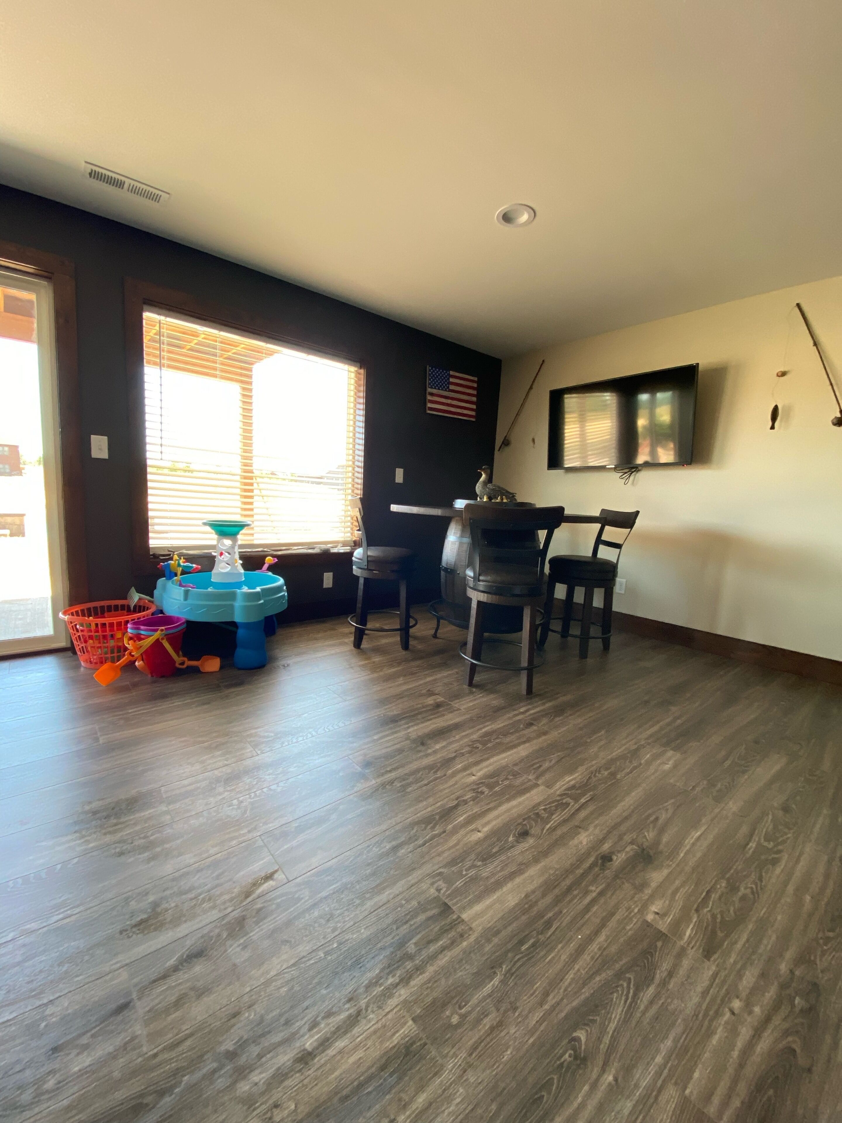 A room with a wooden floor, a dining table with four chairs, a TV mounted on the wall, and a window with blinds.