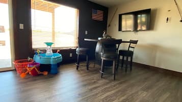 A room with a wooden floor, a dining table with four chairs, a TV mounted on the wall, and a window with blinds.