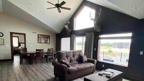 A spacious living room with a brown leather sofa, a coffee table, and a ceiling fan. There is a dining area with a table and chairs in the background.