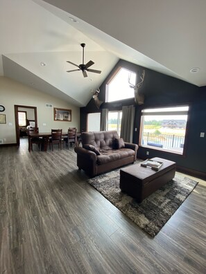 A spacious living room with a brown leather sofa, a coffee table, and a ceiling fan. There is a dining area with a table and chairs in the background. - Large Multifamily Beach Front Lake Home Near WI Dells (Germantown)