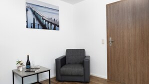 Una habitación con una puerta de madera, un sillón gris, una mesita con una botella y vasos, y un cuadro con una imagen de un muelle.