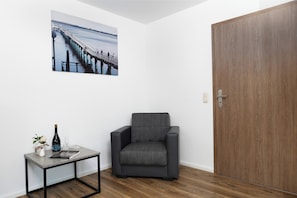 Ein Zimmer mit Holztür, einem grauen Sessel, einem kleinen Tisch mit Flasche und Gläsern sowie einem gerahmten Bild von einem Steg.