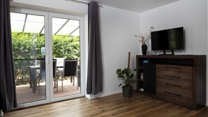 Una habitación con puerta de vidrio corredera que da a un patio exterior, un televisor de pantalla plana sobre un mueble de madera y una planta en maceta.